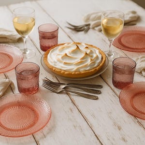 May include: A table set for dessert, featuring pink plates, glasses, and a pie with meringue. Two wine glasses with white wine are visible. Linen napkins and cutlery complete the arrangement, creating a welcoming scene.