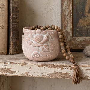 May include: A light pink ceramic planter featuring a raised floral relief design on the front. A strand of brown wooden beads with a small tan tassel is draped over the rim of the planter, resting against the textured surface of the pot.
