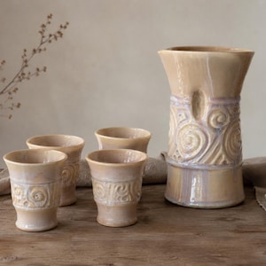 May include: A ceramic pitcher and three matching cups in a glossy beige glaze with iridescent lavender accents. Each piece features a raised, swirling relief pattern around the center, a flared rim, and a smooth, tapered base design.
