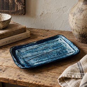 May include: A rectangular ceramic tray featuring a deep blue glaze with a textured, mottled white finish. The surface of the tray has several horizontal grooves running across its length, and the edges are slightly raised and uneven in shape.