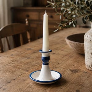 May include: A white ceramic candlestick holder with a flared base and a central stem, featuring dark blue painted rims and horizontal bands. The holder supports a single, slender, off-white taper candle that stands upright in the centre.