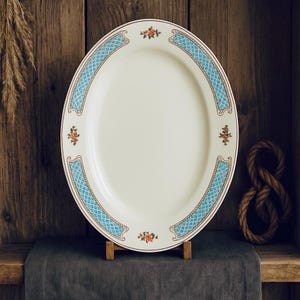 Puede incluir: Una fuente ovalada de color crema con un borde decorativo azul y dorado. El borde presenta un patrón de diamantes y detalles florales. La fuente se exhibe sobre un soporte de madera contra un fondo de madera rústica.