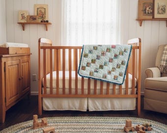 Op de afbeelding: Een licht houten wieg met een blauw-wit quilt en een witte wiegrok. Een houten commode en planken met ingelijste foto's zijn zichtbaar. Een gevlochten ovaal tapijt in blauw, groen en wit ligt op de vloer, samen met houten speelgoedblokken.