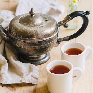 Könnte beinhalten: Eine silberne Teekanne mit schwarzem Griff und zwei weißen Tassen, die mit Tee gefüllt sind. Die Teekanne steht auf einer Holzoberfläche mit einem Leinentuch und einer kleinen Vase mit Gänseblümchen.