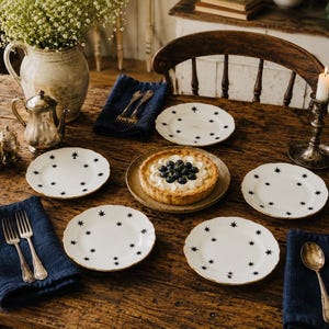 May include: A wooden table displays white plates with black star patterns and gold edges. A blueberry tart is centrally placed, with silverware, blue napkins, and a candle holder. A vase of flowers and a silver tea set complete the scene, creating a charming dining setting.