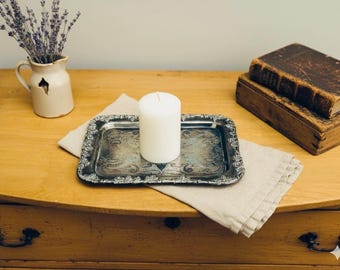 Puede incluir: Una vela blanca sobre una bandeja plateada decorativa con un borde adornado, sobre un paño de lino beige. Un pequeño jarrón con lavanda y dos libros antiguos también están presentes.