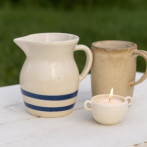May include: A cream ceramic jug with blue stripes, a tan mug, and a small candle holder with a lit candle. The items are arranged on a white surface, suggesting a rustic or farmhouse theme.