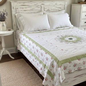 Könnte beinhalten: Ein weißer, abgenutzter Bettrahmen mit passendem Kopfteil. Das Bett ist mit weißer Bettwäsche und einer floralen Steppdecke mit grünen und rosa Akzenten bezogen. Ein kleiner Jute-Teppich liegt auf dem Boden.