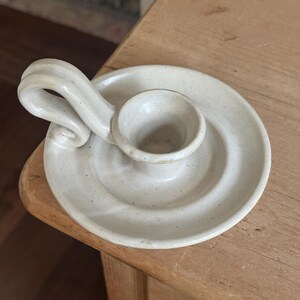 May include: A handmade, off-white ceramic candle holder. The candle holder has a circular base with a raised center for the candle and a curved handle. The piece has a speckled texture and a rustic aesthetic.