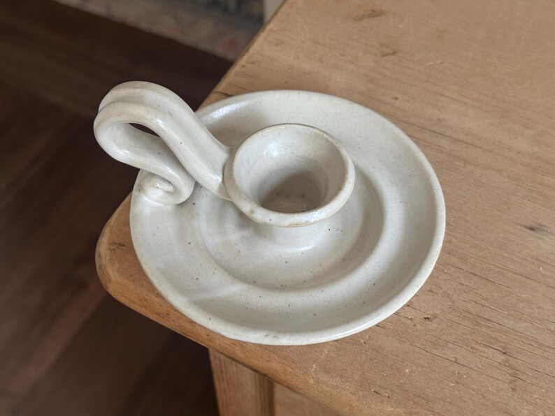 May include: A handmade, off-white ceramic candle holder. The candle holder has a circular base with a raised center for the candle and a curved handle. The piece has a speckled texture and a rustic aesthetic.