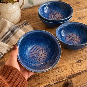 Peut inclure: Trois bols en céramique ronds présentant un émail bleu profond avec un motif de feuilles sculpté en relief à l'intérieur. Les bols ont une finition lisse et brillante avec un bord légèrement plus foncé, révélant un aspect artisanal nuancé.