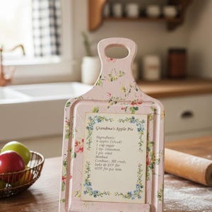 May include: A pink, floral-patterned chopping board with a handle, displaying a recipe for "Grandma's Apple Pie." The recipe card is framed within the board. Apples in a basket and a rolling pin are nearby.