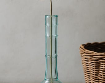 May include: A clear, light blue glass vase with a round base and a cylindrical neck, holding a single, fluffy, beige flower. The vase sits on a wooden surface, with a wicker basket and a book in the background.