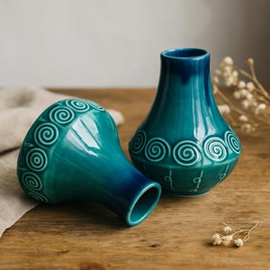 May include: Two teal ceramic vases with a glossy finish. Each vase features a rounded body and a narrow neck. The vases are decorated with a repeating spiral pattern around the body. One vase is upright, and the other is lying on its side.