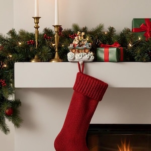 May include: A red knitted Christmas stocking hangs from a white mantelpiece decorated with festive greenery, candles, and gifts. A small decorative train sits on the mantel, adding to the holiday theme.