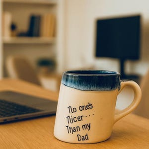 May include: A beige ceramic mug with a blue rim and handle, on a wooden desk beside a laptop. The mug features the text "No one's Nicer... Than my Dad" in black script. A computer monitor and bookshelf are in the background.