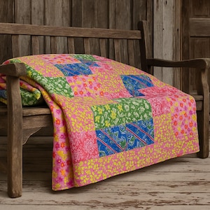 Peut inclure: Une courtepointe colorée à motifs de patchwork est posée sur un banc en bois. La courtepointe présente des carrés de tissu rose, vert et bleu avec des motifs floraux et à motifs. Le banc est en bois vieilli.
