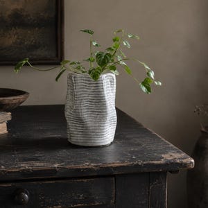 Peut inclure: Un vase cylindrique en céramique avec une surface texturée et ondulée dans des tons blanc cassé et gris. Le vase présente une forme distincte, irrégulière et façonnée à la main, contenant une petite plante verte aux feuilles délicates.