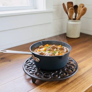 Puede incluir: Una olla humeante de estofado se encuentra sobre un salvamanteles decorativo de hierro fundido. La olla negra tiene un asa plateada y contiene trozos de carne y verduras. Cucharas de madera descansan en un tarro de cerámica. La escena está en una encimera de madera.