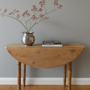 May include: A wooden drop-leaf table with turned legs. A silver vase holds branches with red berries. Two books are stacked on the table. The table is set against a gray wall and a wood floor.