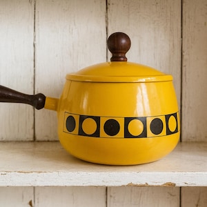 Op de afbeelding: Een gele emaille steelpan met een deksel en een lange handgreep. De pot heeft een zwart-geel geometrisch ontwerp rond het midden. De handgreep en de knop van het deksel zijn donkerbruin.