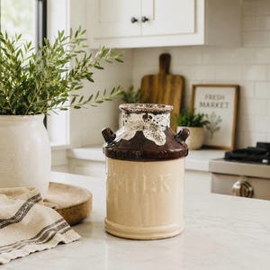 Peut inclure: Pichet à lait en céramique crème avec un dessus et des poignées marron foncé. Le mot "MILK" est imprimé sur le devant. Le pichet est posé sur un comptoir blanc dans une cuisine.