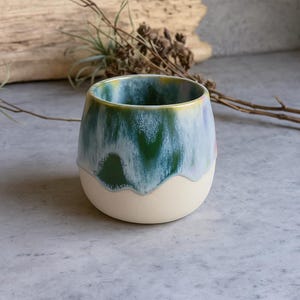 May include: A handmade ceramic tumbler with a unique glaze. The top half features a blend of green and blue hues, while the bottom half is a creamy off-white. The rim has a touch of yellow. This artful piece is perfect for beverages.