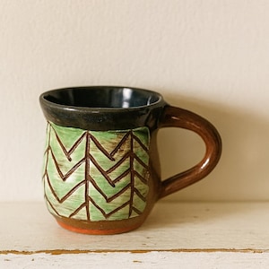 Peut inclure: Une tasse en céramique faite à la main avec une anse et une base marron, un bord noir et un motif en zigzag vert et marron. La tasse est posée sur une surface blanche.