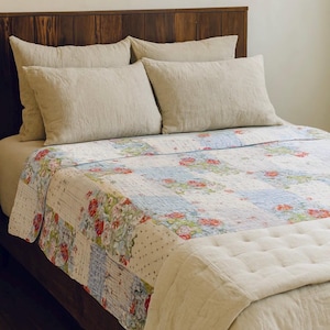 May include: A bed with a wooden headboard and beige bedding. The bed is covered with a patchwork quilt featuring floral patterns in red, blue, and green. Beige pillows are arranged on top of the bed.