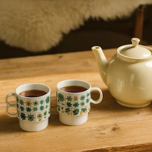 May include: Two white ceramic mugs with green and yellow floral designs, filled with tea, sit on a wooden table next to a matching teapot. A stack of books is also on the table. The teapot and mugs are a cream color.