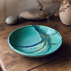 Peut inclure: Un bol en céramique turquoise avec des motifs bleus et verts, posé sur une table en bois. Le bol présente un motif unique et organique. Une petite pierre et un vase sont en arrière-plan. Fabrication artisanale.
