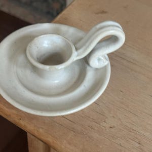 May include: Off-white ceramic candle holder with a circular base and a small cup to hold the candle. It has a curved handle on the side. The candle holder is sitting on a wooden surface.