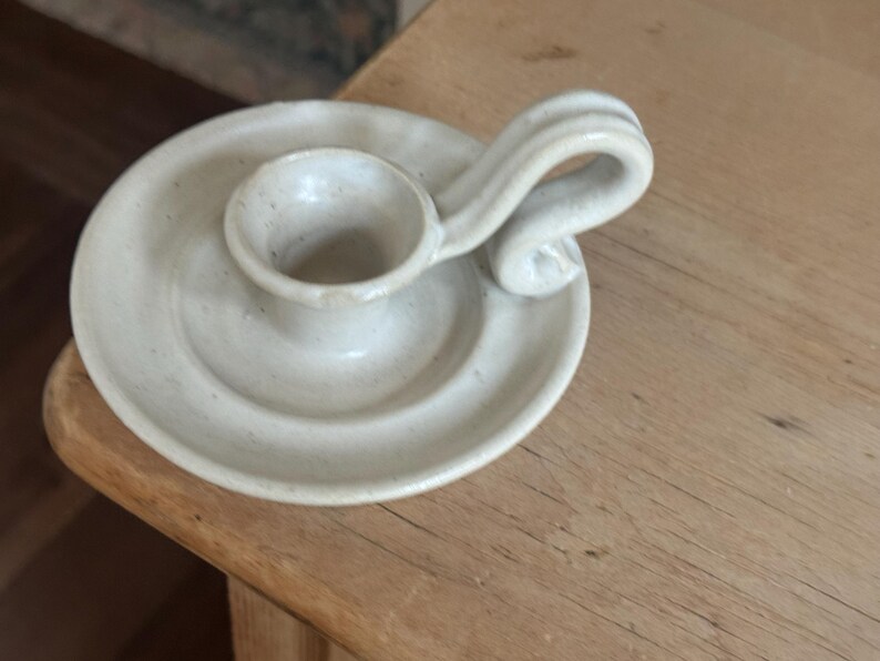May include: Off-white ceramic candle holder with a circular base and a small cup to hold the candle. It has a curved handle on the side. The candle holder is sitting on a wooden surface.