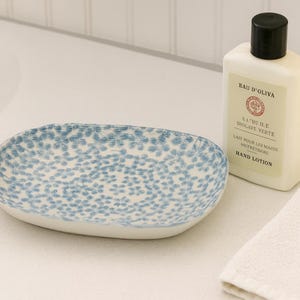 May include: A blue and white floral patterned soap dish, a bottle of hand lotion, and a white washcloth are arranged on a surface. The lotion bottle has a black cap and the text "Eau D'Oliva".