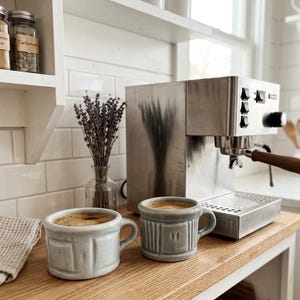 以下が含まれることがあります： 木製のハンドルが付いたステンレス鋼のエスプレッソマシンが木製のカウンタートップに置かれています。コーヒーが入った2つの水色のマグカップがマシンの前にあります。ラベンダーの小さな花瓶とスパイスジャーが棚にあります。