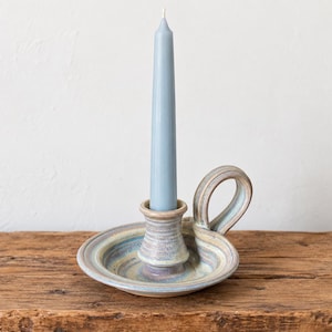 May include: A ceramic candle holder with a circular base and a side handle, featuring a multi-coloured glaze in shades of blue, tan, and cream. A single light blue taper candle is inserted into the central socket of the holder.