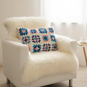 May include: A white armchair with a cream-colored faux fur throw and a crocheted pillow. The pillow features a square pattern in shades of blue, pink, and cream. The chair has silver legs and is placed in front of a window.