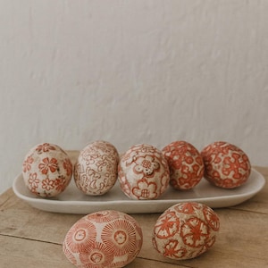 May include: A collection of decorative eggs with intricate red floral patterns on a white background. The eggs are displayed on a white oval platter and a wooden surface. The eggs are a cream color with red floral designs.