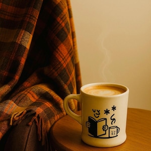 May include: A beige ceramic mug with a cartoon illustration of a person reading a book and a smaller mug. The mug is filled with a latte, and steam rises from the top. A plaid blanket in shades of orange and brown is draped over a chair.