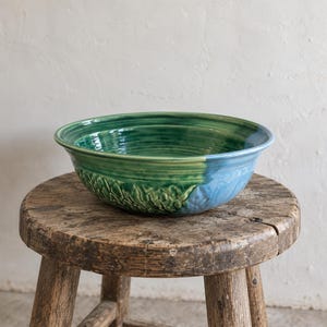 Peut inclure: Un bol en céramique avec une finition brillante présentant un dégradé de couleurs passant du vert profond au bleu clair. La surface extérieure affiche un motif texturé en forme de feuille sur la partie verte, tandis que l'intérieur est lisse.