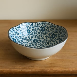 May include: A white ceramic bowl with a scalloped edge and a blue floral pattern. The interior has a detailed design of small flowers and leaves. The rim is outlined with a thin grey border. The bowl is on a wooden surface.