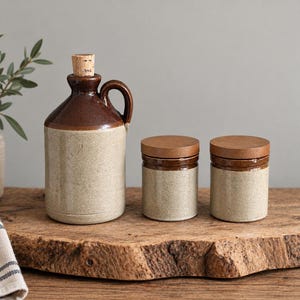 Könnte beinhalten: Ein Keramikkrug mit Korkstopfen und zwei passende zylindrische Vorratsdosen mit Holzdeckeln. Die Gefäße haben ein zweifarbiges Design mit einem dunkelbraun glasierten oberen Teil und einem gesprenkelten, hellbeigen unteren Bereich.