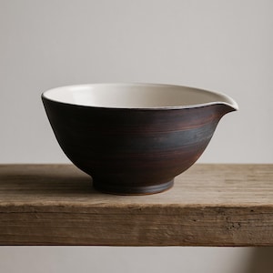 May include: A dark brown ceramic mixing bowl with a white interior. The bowl has a spout and sits on a wooden surface. The exterior has a textured, layered appearance, and the rim is smooth.