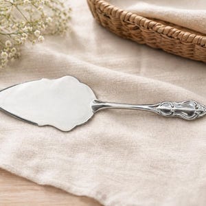 May include: A silver cake server with a decorative handle lies on a beige linen cloth. The server features a wide, scalloped blade and an ornate handle. A woven basket and white flowers are in the background.