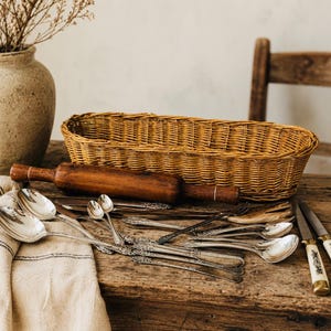 May include: A collection of silver-toned metal spoons and forks with ornate handles, two knives with cream-colored handles, a wooden rolling pin, and an oval woven wicker basket arranged on a rustic wooden surface with a beige linen cloth.