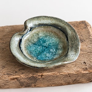 Peut inclure: Un petit plat en céramique en forme de cœur irrégulier avec un bord extérieur moucheté gris et brun. Le centre du plat présente un bassin texturé et cristallin de verre bleu vif, créant un contraste avec le bord en céramique terreuse.
