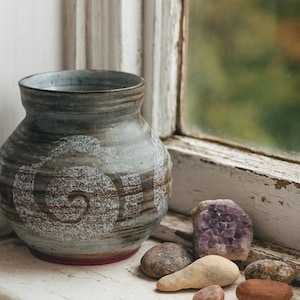 May include: A handmade ceramic vase with a spiral pattern in grey and brown tones, set on a white windowsill. Beside the vase are various small stones and a piece of amethyst. The scene is illuminated by natural light.