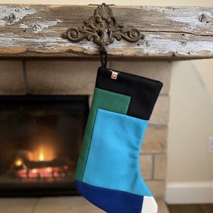 Puede incluir: Un calcetín navideño colorido con un diseño geométrico, colgado de un gancho de metal decorativo. El calcetín presenta paneles negros, verdes, azules y azul marino, con una punta blanca. Una chimenea acogedora es visible en el fondo.