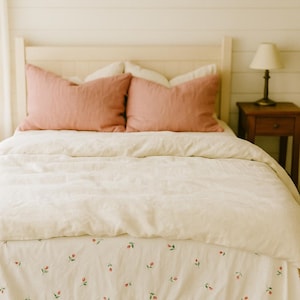 Pode incluir: Uma cama com cabeceira creme e roupa de cama a condizer. Duas almofadas cor de rosa empoeirado repousam contra a cabeceira. A saia da cama apresenta um delicado padrão floral com detalhes em vermelho e verde. Uma pequena mesa de cabeceira de madeira com uma lâmpada está à direita.