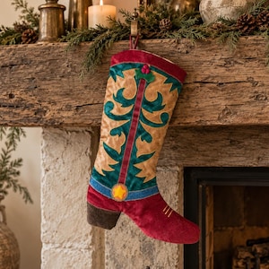 Peut inclure: Une chaussette de Noël en forme de botte de cow-boy, avec un pied et un revers en velours rouge foncé, une tige beige avec des superpositions décoratives bleu sarcelle, un emblème en étoile jaune et un motif de houx vert avec des baies rouges.